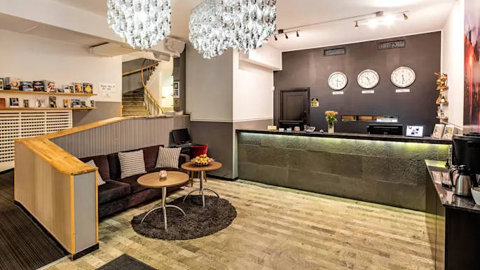 Elegant hotellresepsjon med to lysekroner, resepsjonsskranke, sitteområde, og dekorative klokker på veggen. Moderne design med en varm atmosfære.