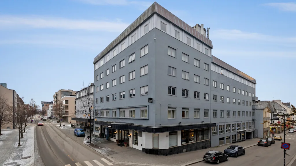 En stor, grå byggnad förankrar ett stadsgathörn, med bilar som rör sig och parkerar, och kala träd under en ljus himmel.