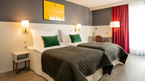 Två prydligt bäddade enkelsängar med gröna kuddar och grå täcken står i ett modernt hotellrum. Gul konst med texten "BANANA" hänger ovanför; ett skrivbord och stol syns nära röda gardiner.