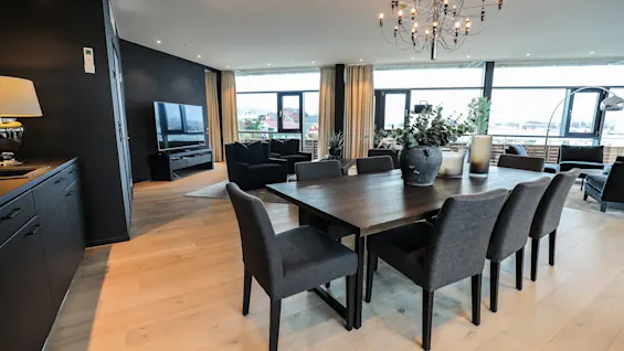 Et romslig spisestue med et mørkt trebord og åtte stoler står under en lysekrone i en elegant innredet, moderne interiør med store vinduer og en TV.