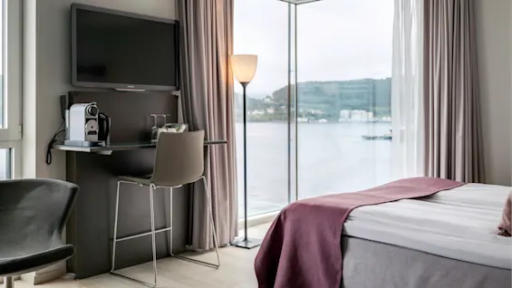 Ett modernt hotellrum med en säng med lila detaljer, en väggmonterad TV, en stol vid ett skrivbord med en kaffemaskin och stora fönster med utsikt över vatten.