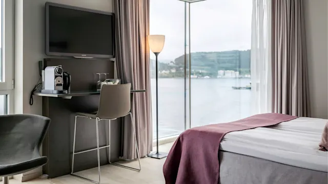 A modern hotel room features a bed with purple accents, a wall-mounted TV, a chair at a desk with a coffee machine, and large windows overlooking a tranquil waterside view.