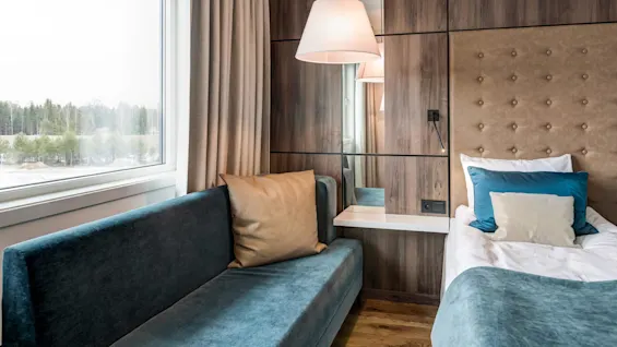 A cozy room features a modern sofa and bed with cushions beside a large window displaying a forest view. Warm wooden panels and soft lighting enhance the inviting atmosphere.