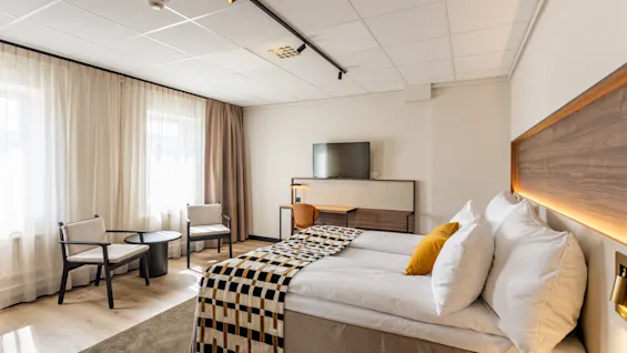 Valoisa ja moderni hotellihuone, jossa kaksi sänkyä, työpöytä, televisio ja viihtyisä oleskelualue ikkunan vieressä.
