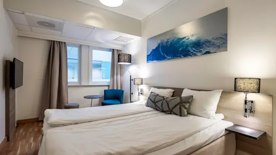 A bed with white linens is positioned in a modern room, flanked by nightstands. A large wave artwork decorates the wall, and a blue chair with a table sits by the window.