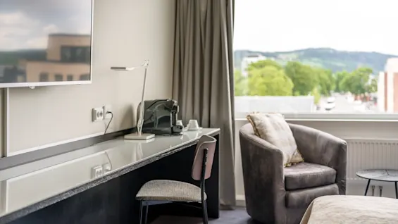 A modern hotel room features a neatly arranged desk with a lamp and chair. A comfy armchair sits near a large window offering a scenic view of greenery and hills.