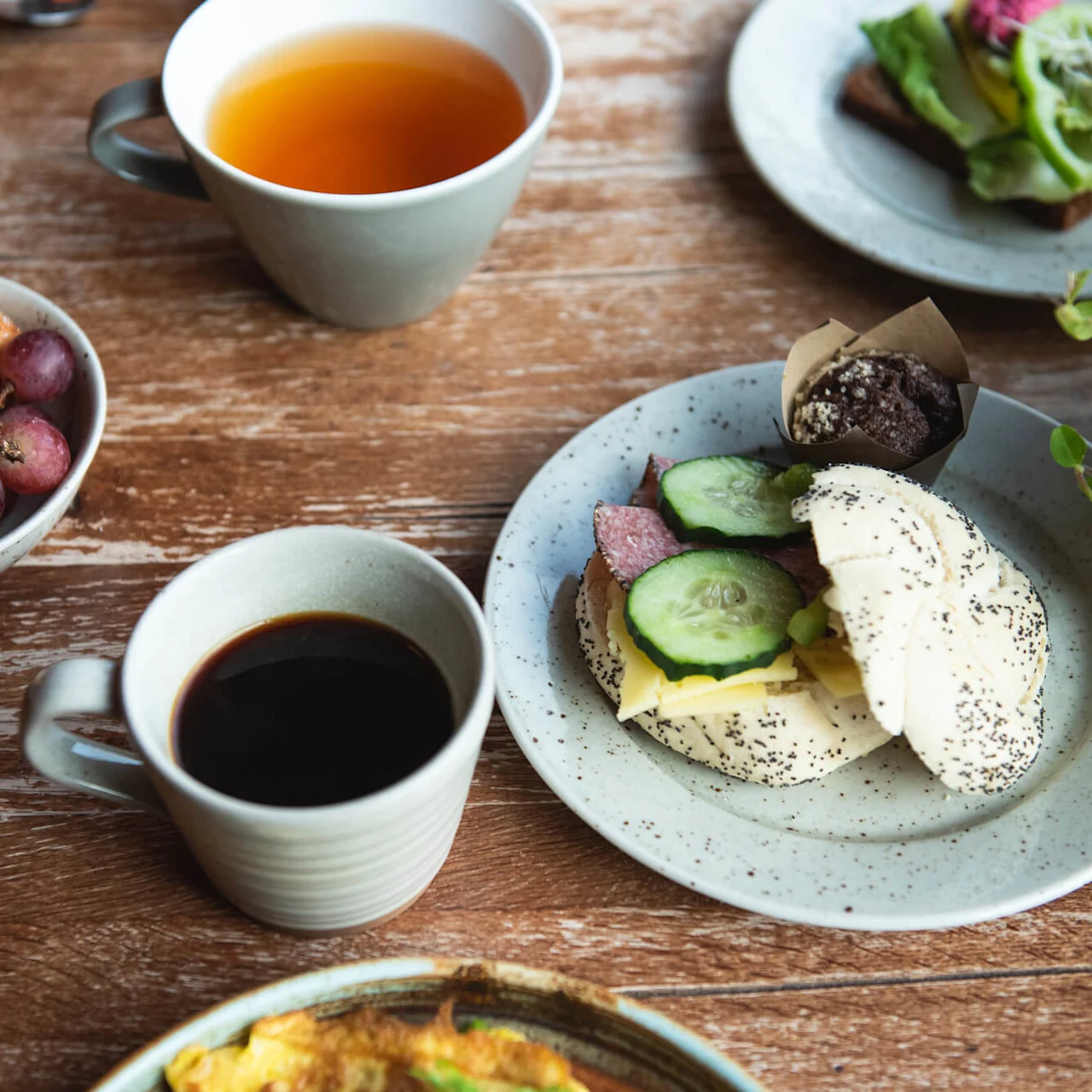 A wooden table holds various breakfast items: a poppy seed sandwich with cucumber and cheese, a muffin, coffee, tea, grapes, a croissant, and fruit slices, complemented by greenery.