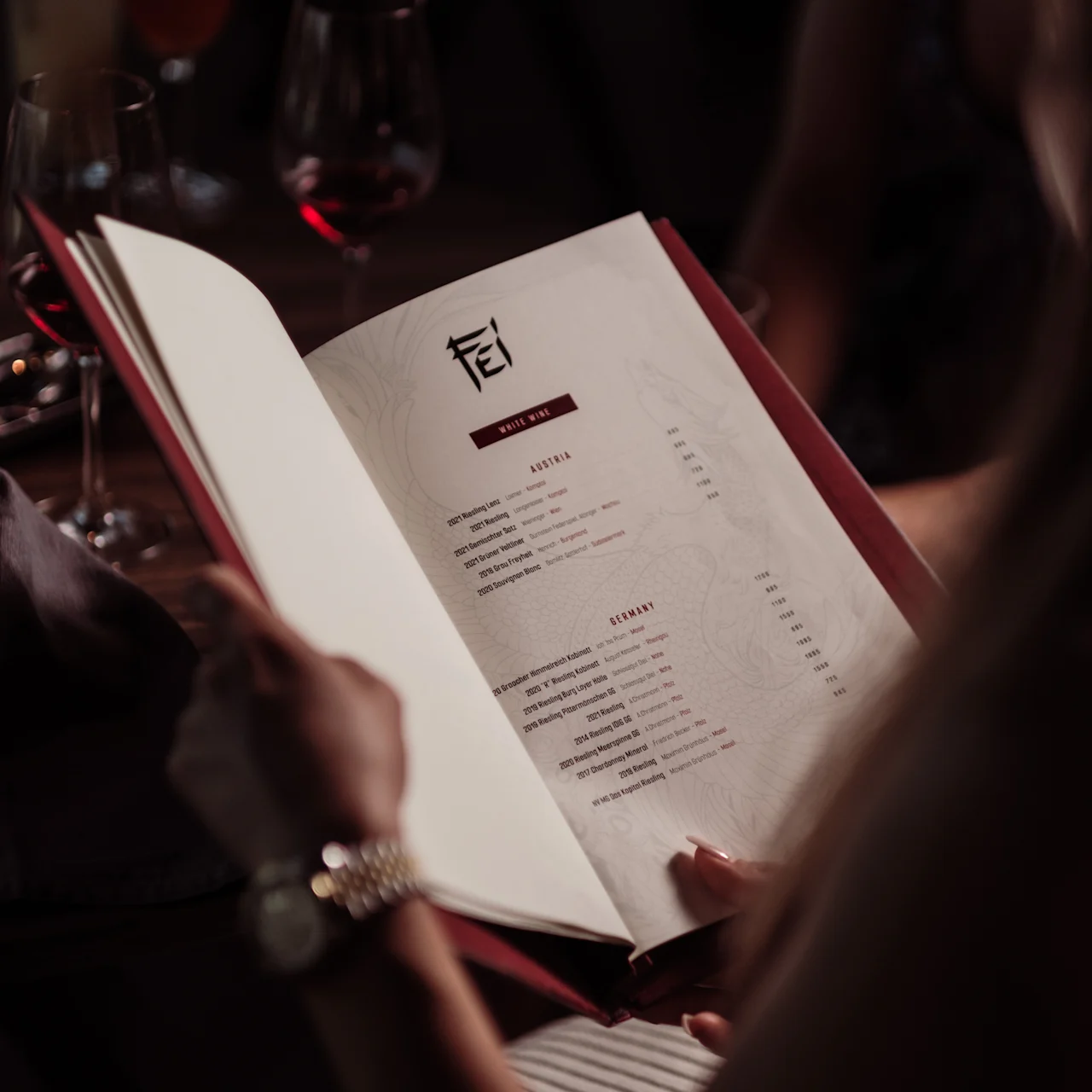 En person håller en restaurangmeny med röd rygg med texten "FEI," med dryck- och maträtter, vid ett svagt upplyst bord med vinglas och en cocktail.