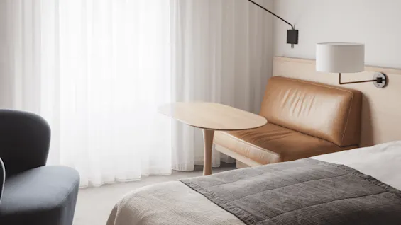 A brown leather bench is adjacent to a compact wooden table, situated beside a bed with a grey quilt, in a room illuminated by soft natural light through sheer curtains.