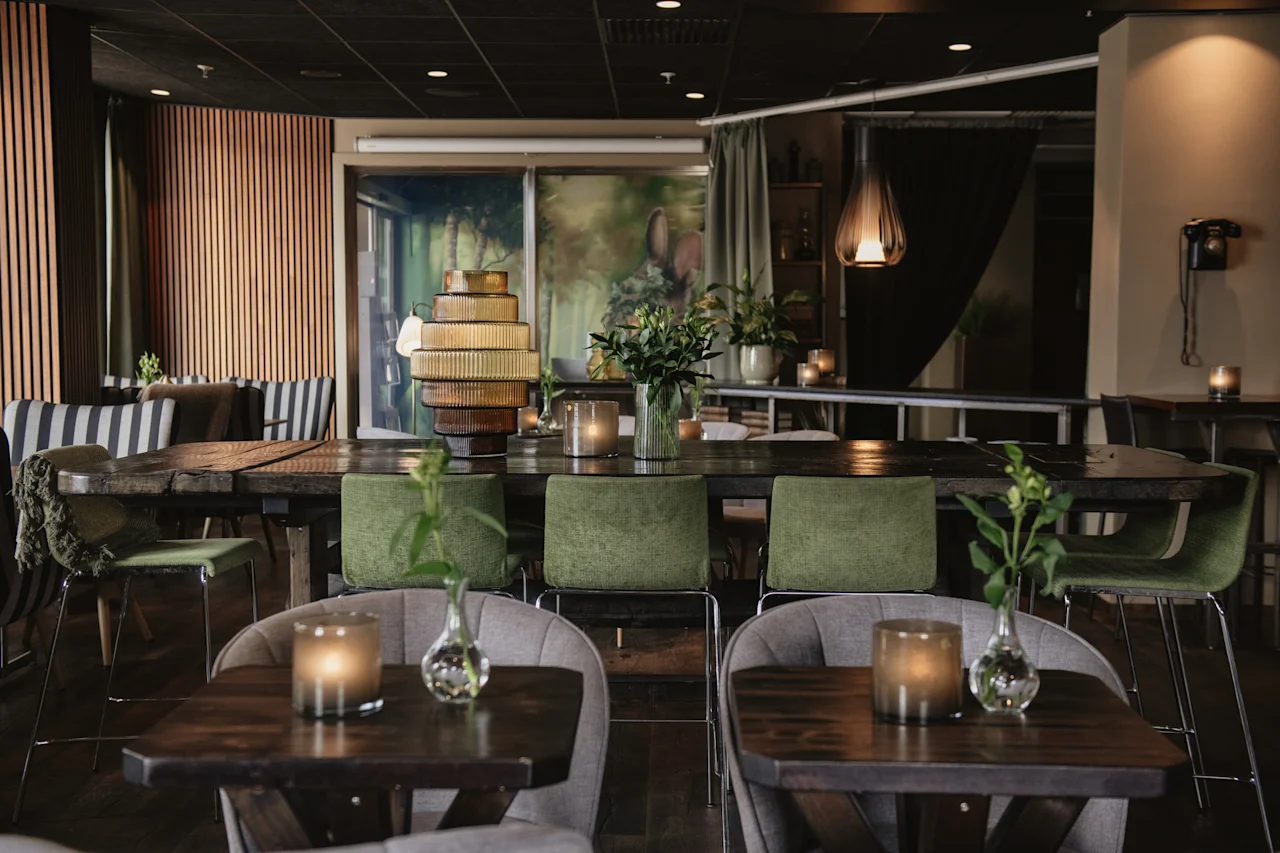 Bord med ljus och vaser står i en svagt upplyst, stilfull restaurang med gröna stolar och modern inredning. Ett stort konstverk pryder väggen och kompletterar den lugna atmosfären.