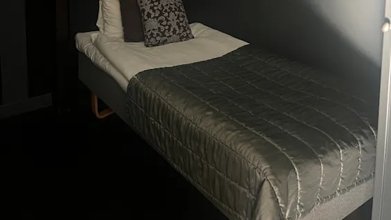 A single bed with a gray quilt, white sheets, and patterned pillows stands against dark striped walls in a dimly lit room.