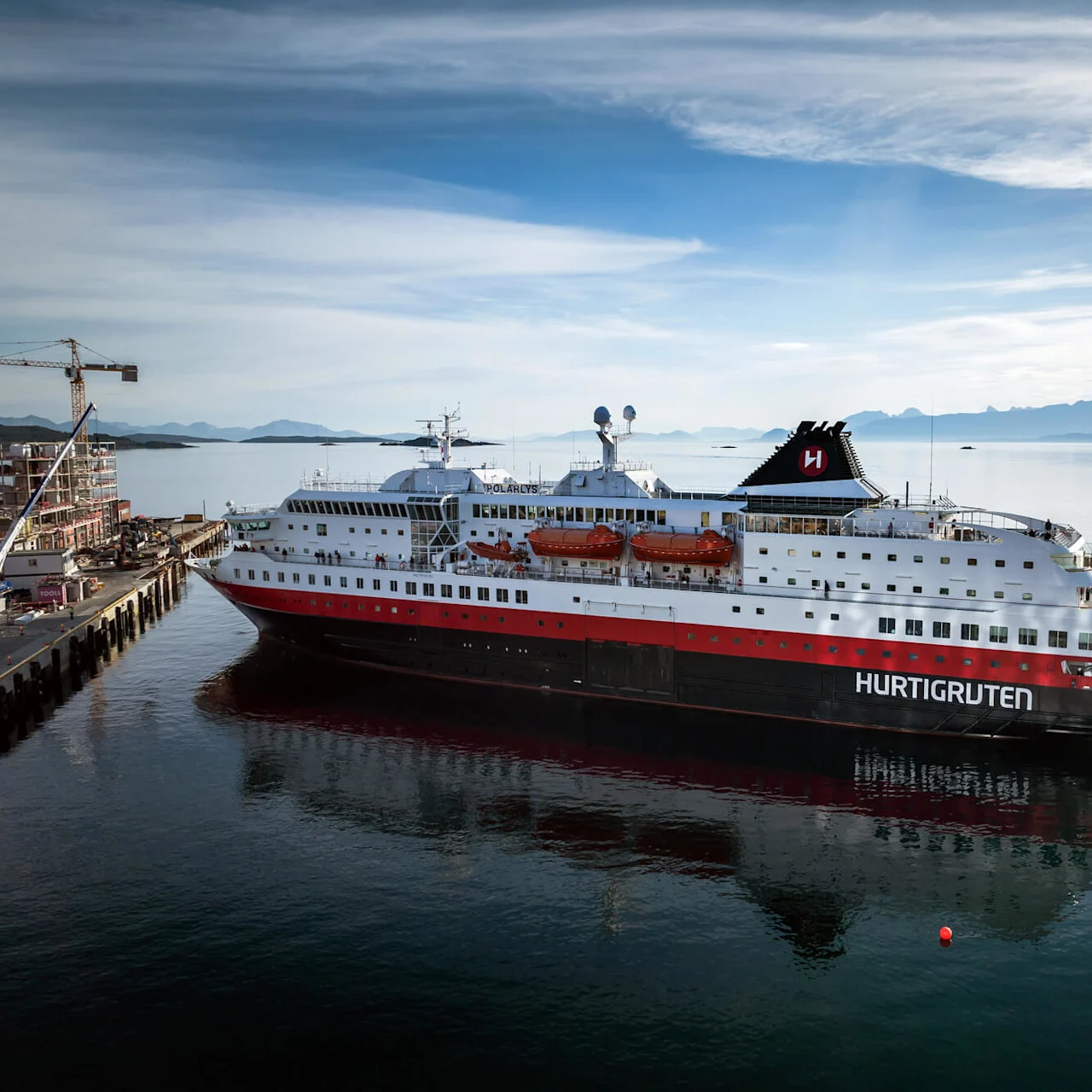 Et stort krydstogtskib mærket "HURTIGRUTEN" og "POLARLYS" sejler gennem rolige vande nær en kaj med en bygningskran, omgivet af fjerne bjerge under en klar himmel.
