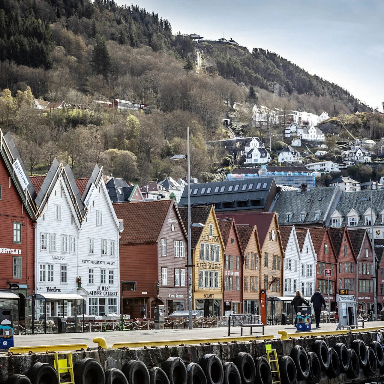 Farverige træhuse langs en kystlinje, omgivet af et bjerglandskab med træer og spredte huse. Tekst inkluderer: MADAM FELLE, BRYGGEN NIGHTCLUB, MARINA, JUHL'S SILVER GALLERY, SCHØTT, D/STØRKEN, M/S BAKEREN.