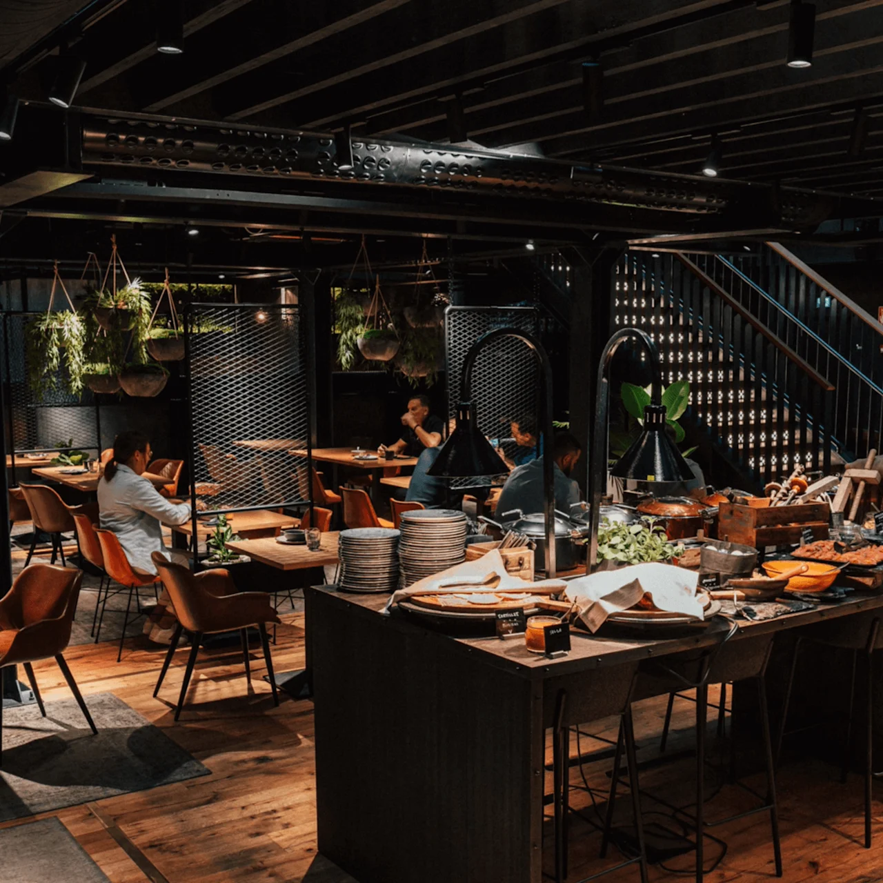 Et buffetbord viser forskjellige retter og tallerkener i en moderne restaurant med tregulv og hengende planter, omgitt av gjester ved bord under svak belysning.