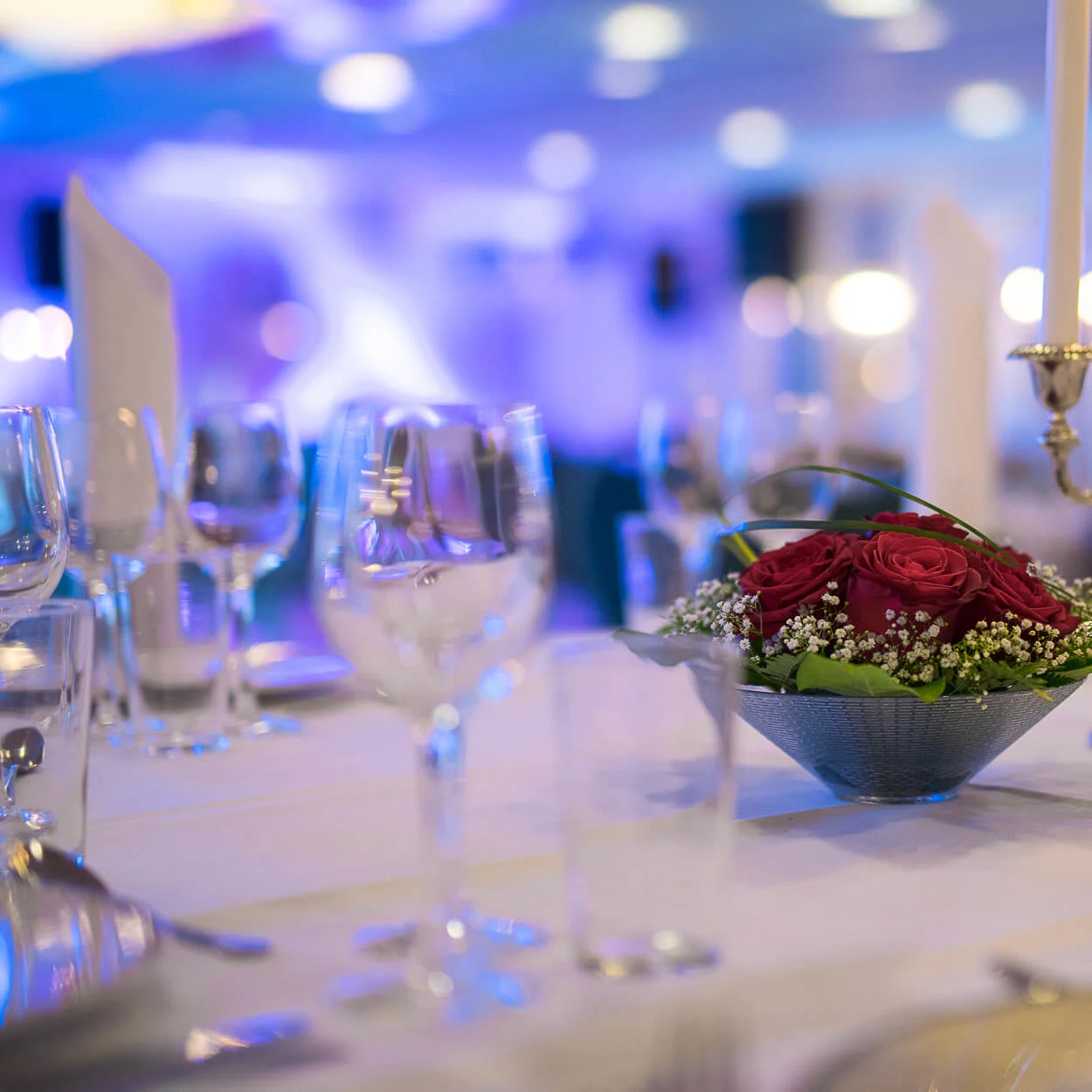 En blomsterdekoration med røde roser og hvide blomster står på et banketbord. Omkring er elegante vinglas, bestik og en lysestage; baggrunden er blødt sløret.