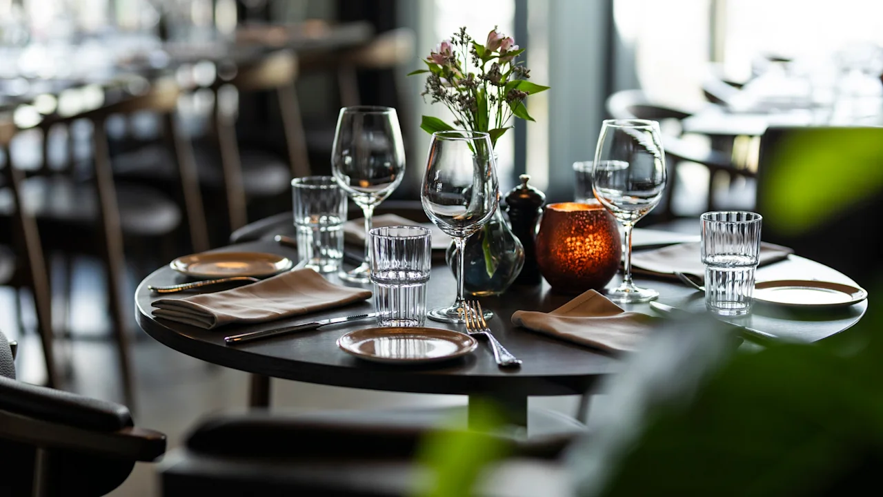 Et smukt dækket restaurantbord med glas, bestik, servietter, blomster og et glødende lys, klar til middag.