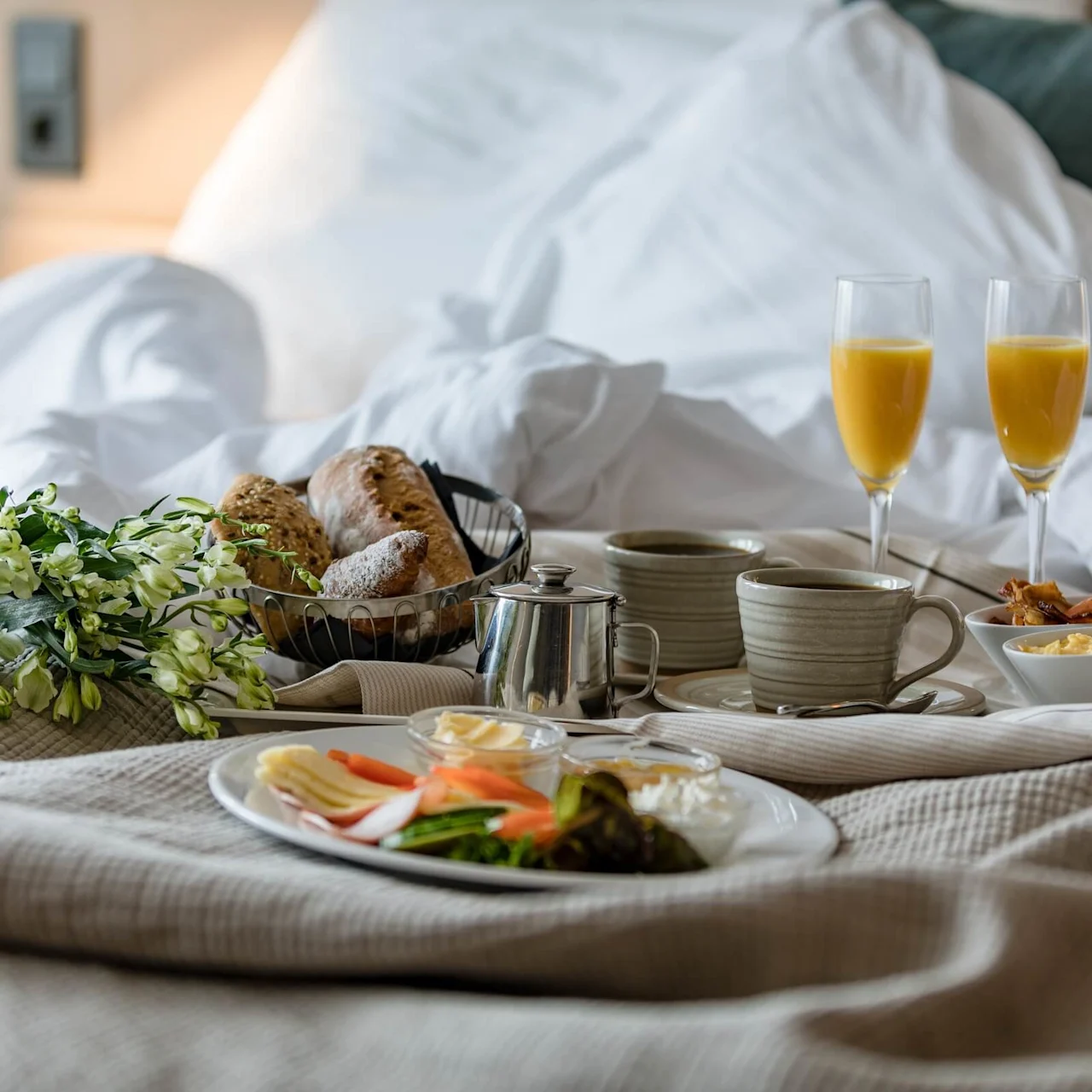Breakfast tray holds croissants, mugs, plates of cheese and salad, glasses of orange juice, and flowers. Set on a bed with white linens.