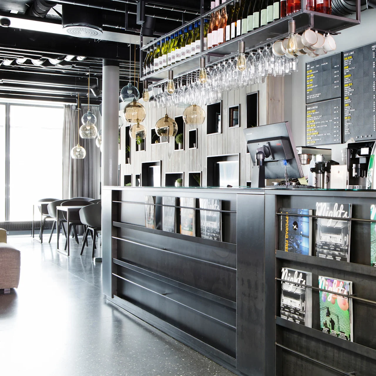 A modern bar counter displays magazines, with bottles and glassware overhead. A menu board on the wall lists drinks. Tables and chairs are arranged in the background. Text: Nights.
