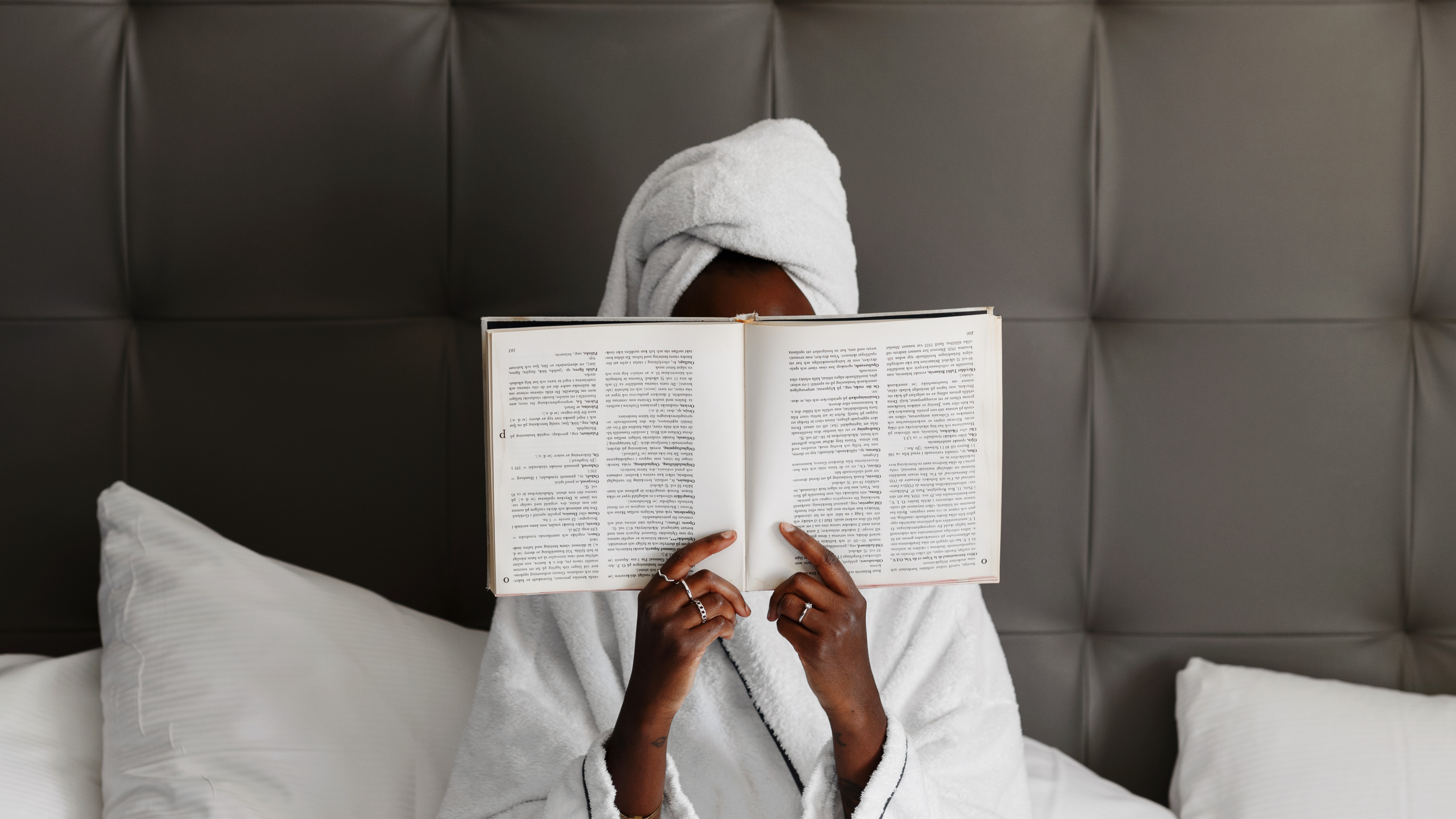 En person i badekåbe og håndklæde på hovedet, slapper af i sengen og læser en bog.