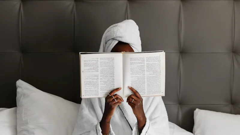 En person i badekåbe og håndklæde på hovedet, slapper af i sengen og læser en bog.