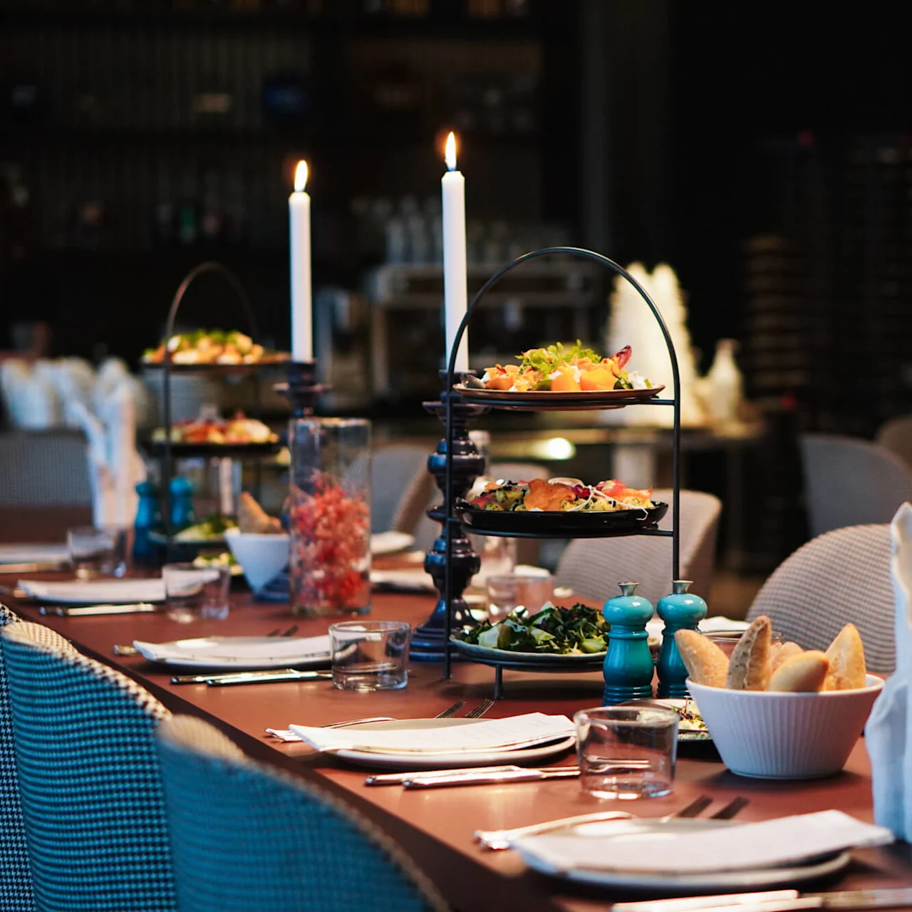 A long dining table holds tiered platters of appetizers, flanked by lit candles and surrounded by elegant place settings with folded napkins in a dimly lit restaurant. 