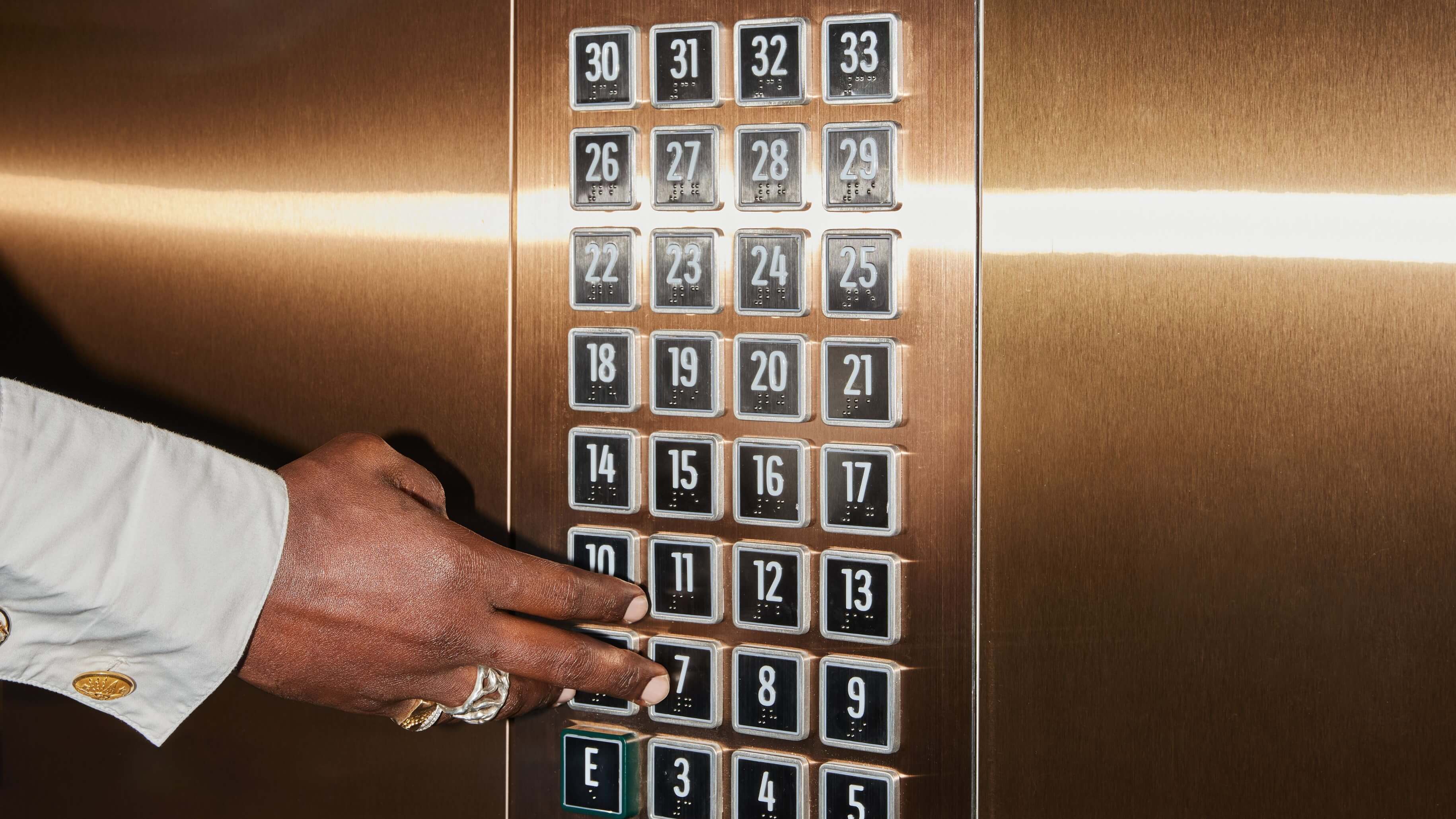 En hånd trykker på en elevator knap, reflekterende overflader er allestedsnærværende, et sekundært mindre billede viser en person der taler i telefon.