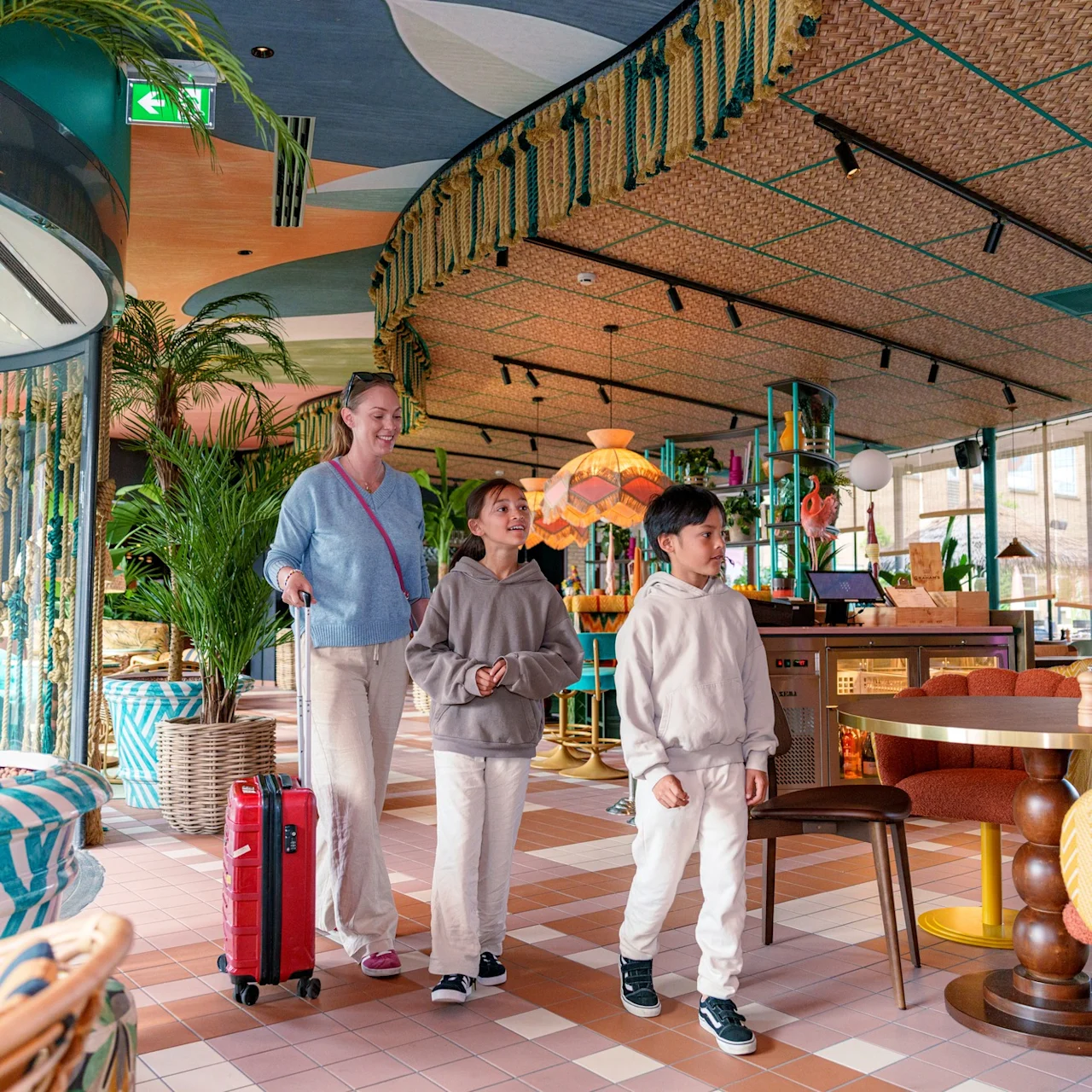 A woman pulls a red suitcase, walking with two children inside a colorful, plant-filled restaurant featuring patterned furniture and decorative ceilings.
