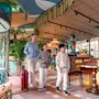 En kvinde trækker en rød kuffert og går med to børn i en farverig, plantefyldt restaurant med mønstrede møbler og dekorative lofter.