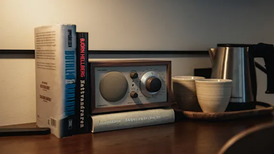 Et hyggeligt arrangement på et træbord med bøger, en radio mærket MODEL ONE, og en tekande med kopper. Elden och askan.