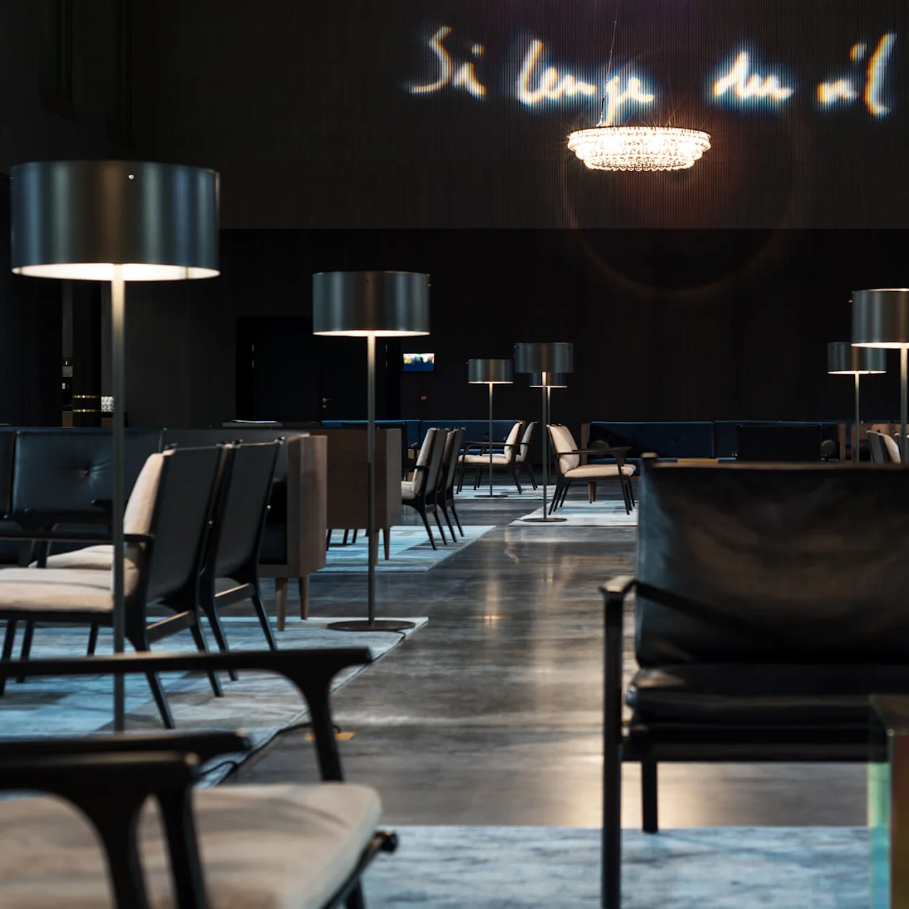 Sofaer og stoler er pent arrangert i en romslig, moderne salong med lamper og en stor lysekrone. Veggen viser teksten "Så lenge du vil."
