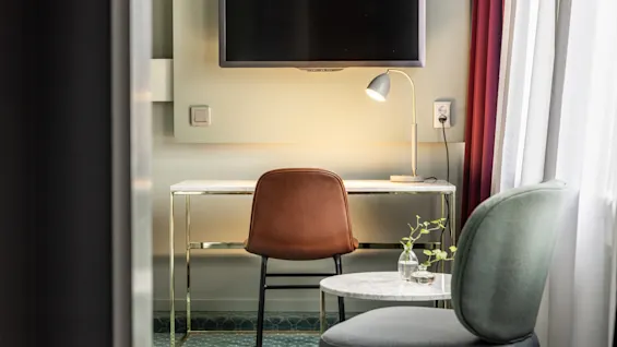 Ett mysigt arbetsutrymme på hotellrummet med ett modernt skrivbord, en lampa, väggmonterad TV och en bekväm stol. Naturligt ljus strömmar in genom gardinerna.