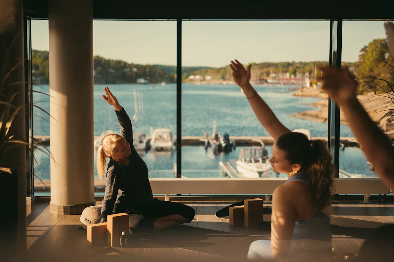 Folk der strækker sig indendørs med en naturskøn udsigt over en sø og både uden for et stort vindue, nyder et fredeligt øjeblik.