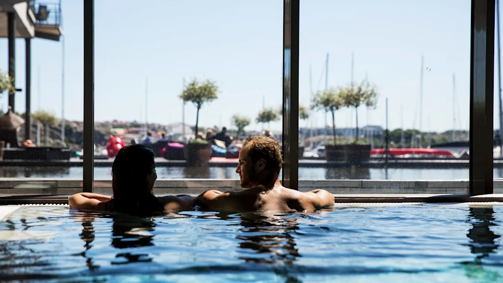 Two people relax in a swimming pool, leaning against the edge. Large windows reveal a marina with moored boats and distant plants in the sunny backdrop.