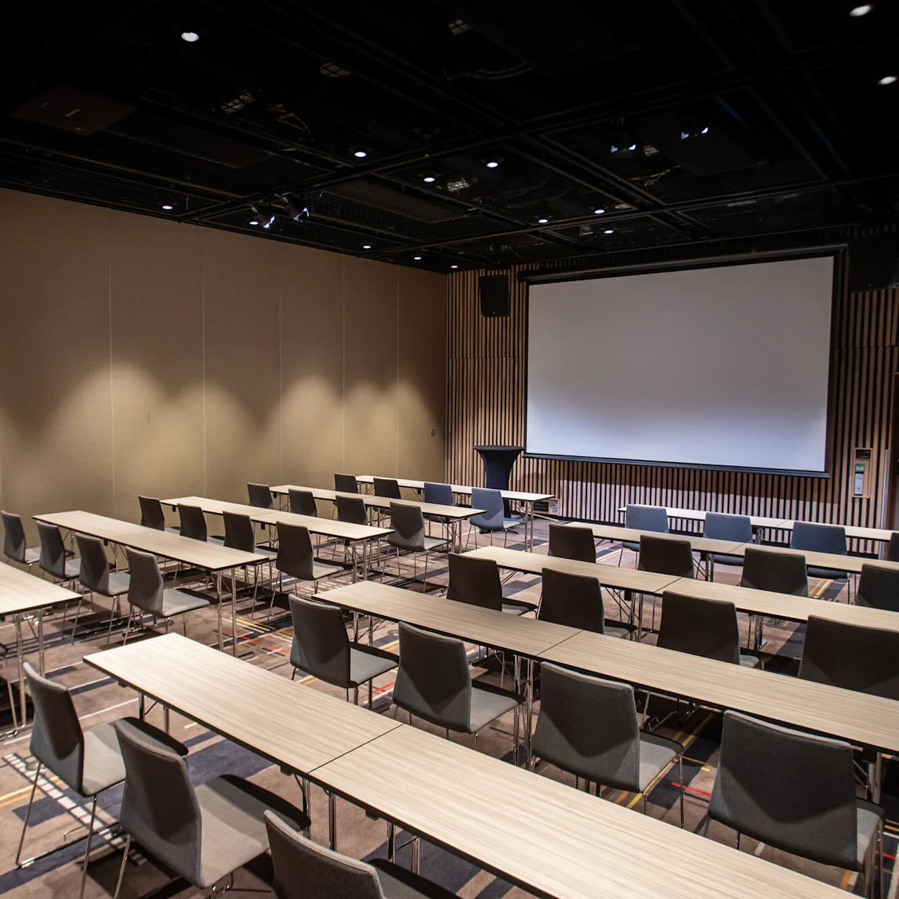 Empty conference room with rows of tables and chairs facing a large projection screen. Dim lighting creates a professional atmosphere. Walls feature wooden paneling; a door is visible to the right.