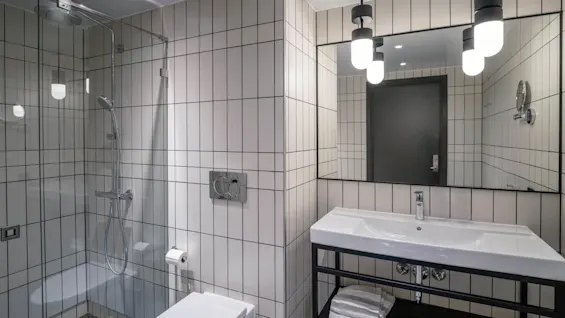 Bad med glasslukket dusj, toalett og bred vask. Geometriske fliser dekker veggene, og tre hengende lamper henger over et stort speil. Håndklær er pent plassert under vasken.