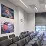 Rows of gray chairs face a wooden desk and a wall-mounted screen in a conference room. Art posters and a wall clock adorn the walls, with light streaming through large windows.