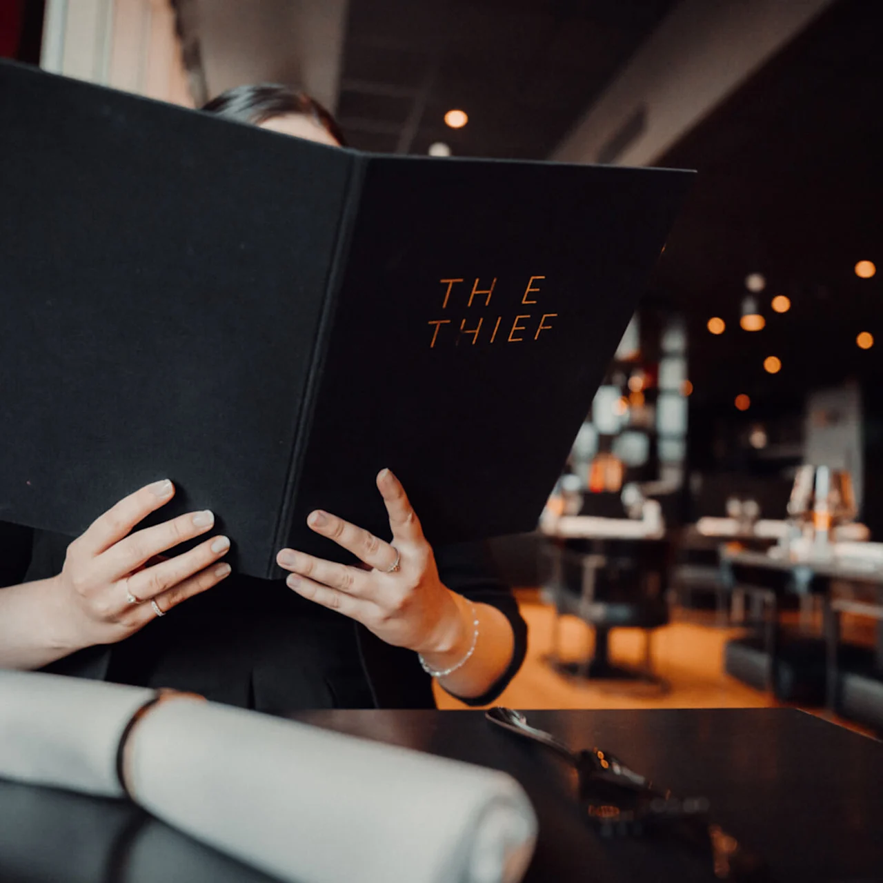 En person holder en stor, sort meny med tittelen "THE THIEF" i en svakbelyst restaurant, omgitt av pent dekkede bord.