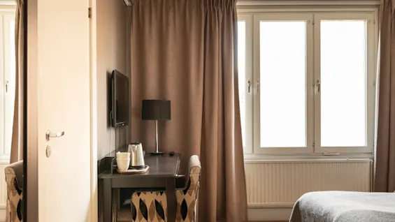 A cozy hotel room with a desk, TV, and coffee amenities, bathed in natural light from a large window with brown curtains.