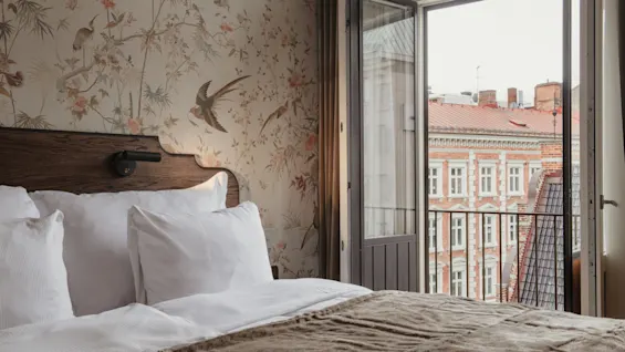 Home Hotel Baltzar - Deluxe Double. Cozy bedroom with bird wallpaper, bed, and open balcony overlooking city rooftops.
