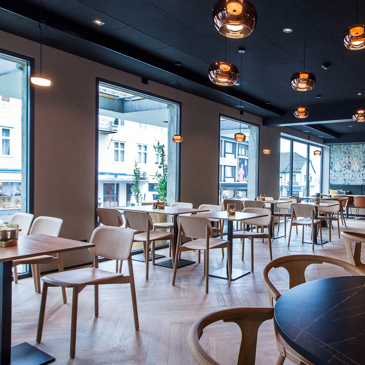 En restaurant med moderne trebord og stoler er tom, opplyst av hengende lamper. Store vinduer slipper inn naturlig lys og vender mot en gate. En dekorativ vegg gir en stilfull touch.