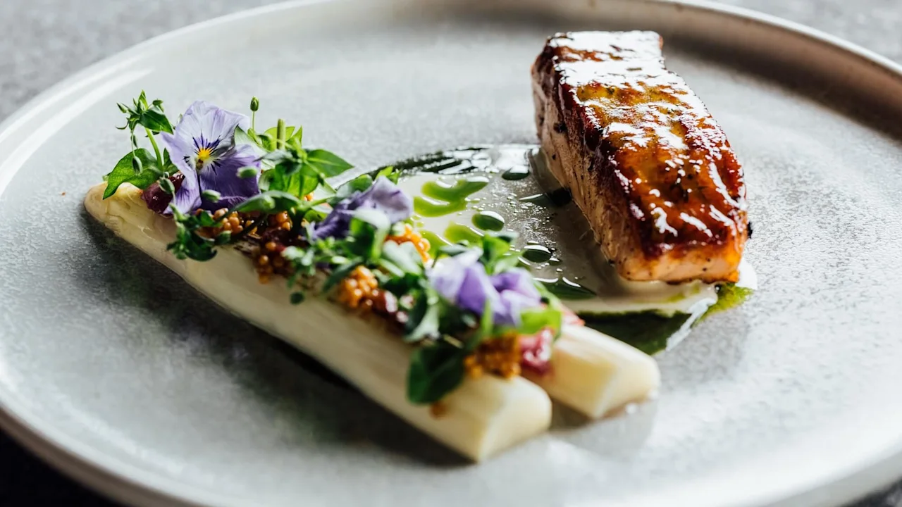 En gourméttallrik med stekt fisk bredvid vit sparris prydd med örter och lila blommor, presenterad på en texturerad grå tallrik i en dämpad belysning.