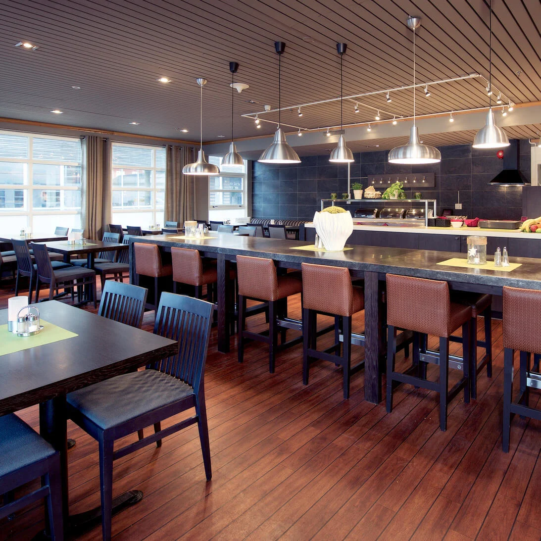Et tomt restaurantlokale med borde, stole og barstole, der viser et moderne åbent køkken på et trægulv under varm belysning.