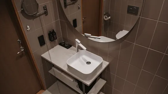 Sink reflecting in a round mirror, surrounded by beige tiles. Hand soap and lotion are beside the sink, with a door slightly open in the background.
