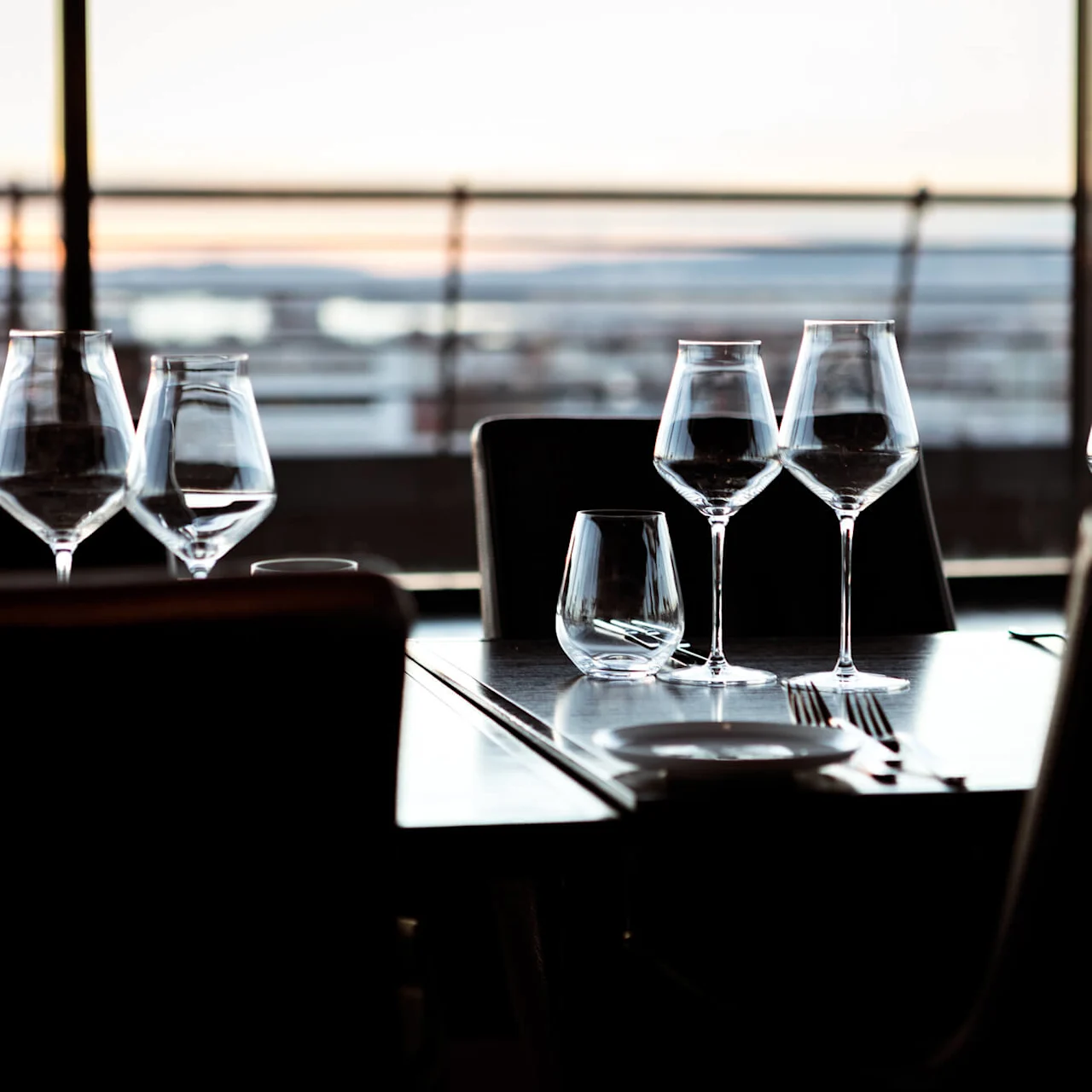 Wine glasses sit on a neatly set table with silverware, overlooking a blurred sunset view through large windows.