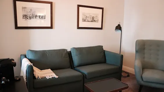 En grønn sofa med et hvitt kast, en matchende lenestol, et sidebord og en gulvlampe, i et koselig rom med innrammede kunstverk på veggene.