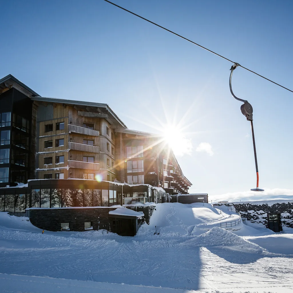 En moderne hotellbygning plassert i et snørikt fjellandskap med en skitrekk over under en lys, solrik himmel.