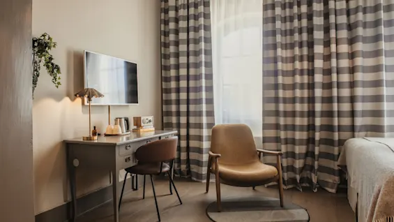 A cozy hotel room featuring a desk with a TV, a comfortable armchair by a window, and striped curtains.