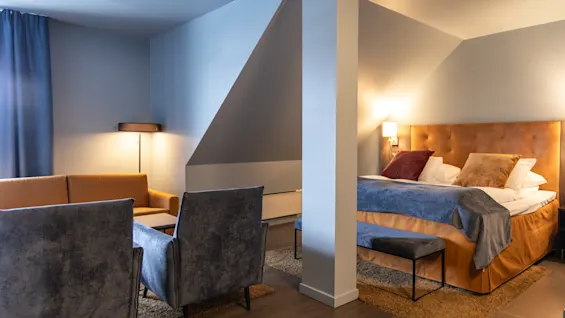 A bed with pillows and a blanket stands in a cozy, dimly lit room, next to armchairs and a low table, under sloped ceilings with soft lamp lighting.