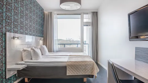 En seng med hvitt sengetøy står mot en mønstret vegg. Nøytrale gardiner rammer inn et vindu. I nærheten dekorere et skrivebord med en stol og TV det minimalistiske rommet.