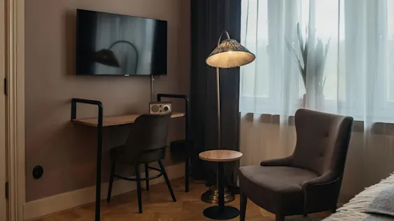 TV mounted on a beige wall, above a wooden desk with a chair and a small radio. Nearby, a floor lamp illuminates a cozy chair next to a window.